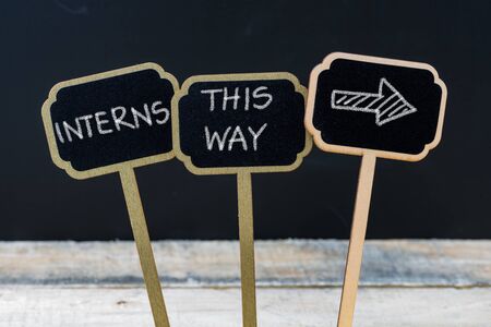 Business message INTERNS THIS WAY written with chalk on wooden mini blackboard labels, defocused chalkboard and wood table in backgroundの写真素材