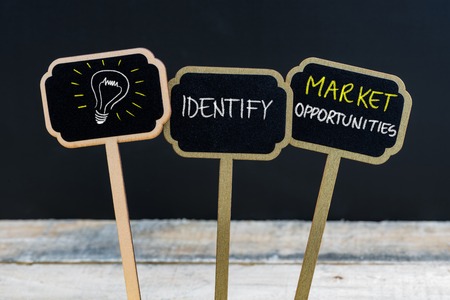 Concept message IDENTIFY MARKET OPPORTUNITIES and light bulb as symbol for idea written with chalk on wooden mini blackboard labels, defocused chalkboard and wood table in backgroundの写真素材