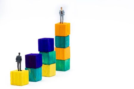 Miniature figurine posed as businessman sitting alone on a wooden cube looking up to leader on colourful stacks as business chart with focus on foreground, minimalist abstract concept imageの写真素材