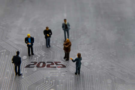 Miniature figurines posed as business people in a meeting over abstract futuristic circuit board with text year 2025の写真素材