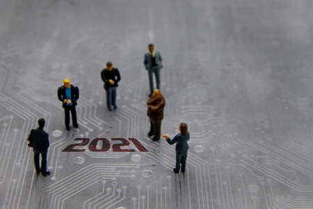 Miniature figurines posed as business people in a meeting over abstract futuristic circuit board with text year 2021の写真素材