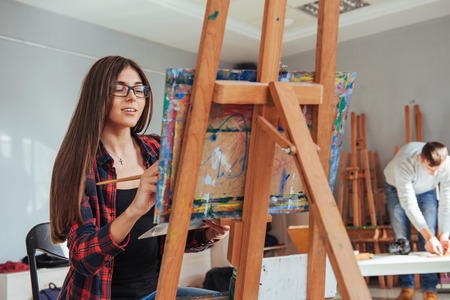 artist painting a picture in a studio. Creative pensive painter girl paints a colorful picture on canvas with oil colors in workshop.の写真素材
