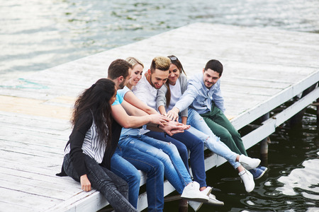 Portrait of joyful group of young people sitting on the edge of the pier, outdoors in nature. Friends enjoying a day on the lake.の写真素材