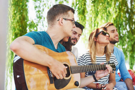 Group of happy friends with guitar. While one of them is playing guitar and others are giving him a round of applauseの写真素材