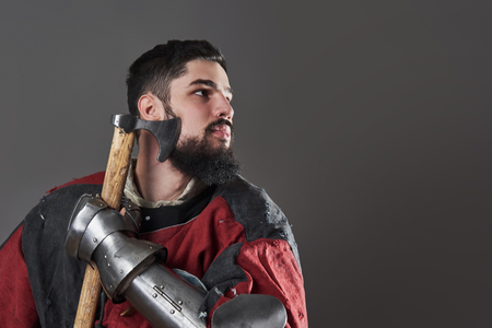 Medieval knight on grey background. Portrait of brutal dirty face warrior with chain mail armour red and black clothes and battle axe.の写真素材