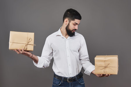 Handsome romantic guy is looking at box and makes a choice. Holding a two big gift box for his couple, on gray background. The concept of choice.の写真素材