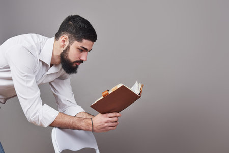 Portrait of a bearded young man wearing a white shirt and holding an open planner and a pen. A gray wall background.の写真素材