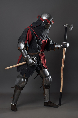 Medieval knight on grey background. Portrait of brutal dirty face warrior with chain mail armour red and black clothes and battle axe.の写真素材