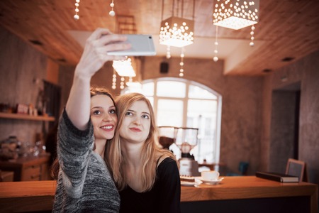 Two friends drinking coffee in a cafe, taking selfies with a smart phone and having fun making funny faces. Focus on the girl on the left.の写真素材