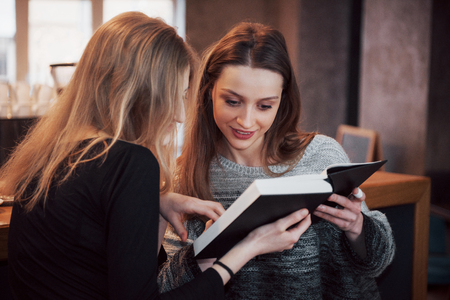Two Girl`s absorbed in reading book during the break in cafe. Cute lovely young women are reading book and drinking coffee.の写真素材
