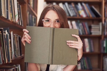 Young attractive student librarian reading a book between library bookshelves.