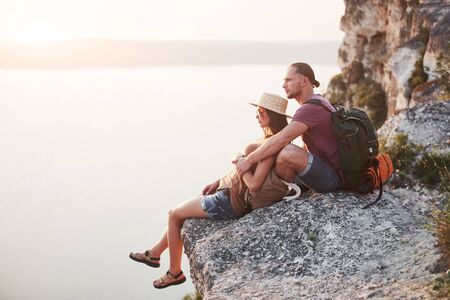 Hugging couple with backpack sitting on top of rock mountain enjoying view coast a river or lake. Traveling mountains and coast, freedom and active lifestyle concept.の写真素材