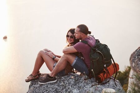 Hugging couple with backpack sitting on top of rock mountain enjoying view coast a river or lake. Traveling mountains and coast, freedom and active lifestyle concept.の写真素材