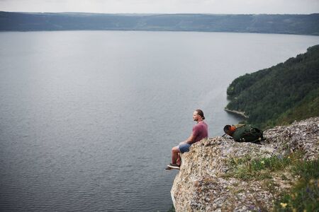 Traveller with backpack sitting on top of mountain enjoying view above the water surface. Traveling along mountains and coast, freedom and active lifestyle concept.の写真素材