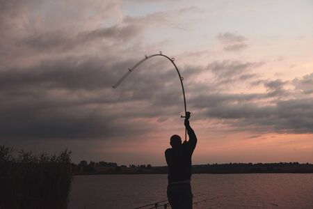 Silhouette of fisherman throws a fishing pole into the lake at sunset. Hunting and hobby sport.の写真素材