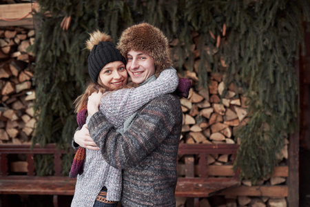 christmas and couple concept - smiling man and woman in hats and scarf hugging over wooden country...