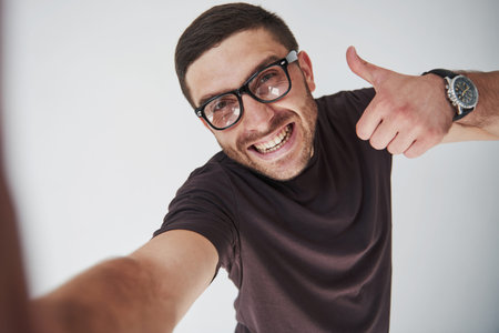 Selfie portrait of a smiling man in glasses showing thumb up over white background.