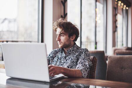 Young handsome man sitting in office with cup of coffee and working on project connected with modern cyber technologies. Businessman with notebook trying to keep deadline in digital marketing sphere.の写真素材