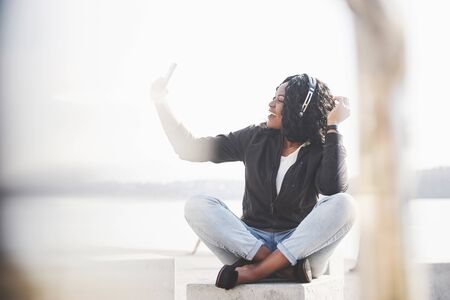 Portrait of a beautiful young pretty African American girl sitting on the beach or lake and listening to music in her headphones.の写真素材