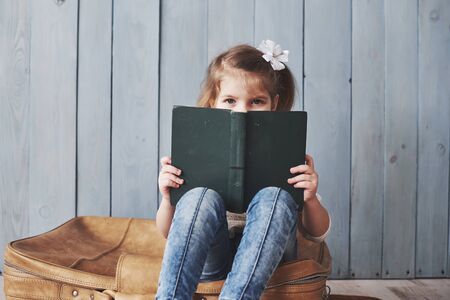 Ready to big travel. Happy little girl reading interesting book carrying a big briefcase. Freedom and imagination concept.の写真素材