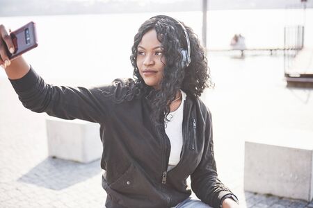 Portrait of a beautiful young pretty African American girl sitting on the beach or lake and listening to music in her headphones.の写真素材