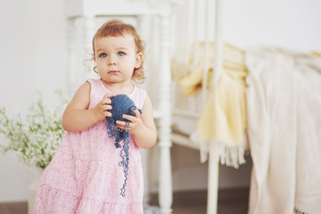 Childhood concept. Baby girl in cute dress play with colored thread. White vintage childroom.の写真素材