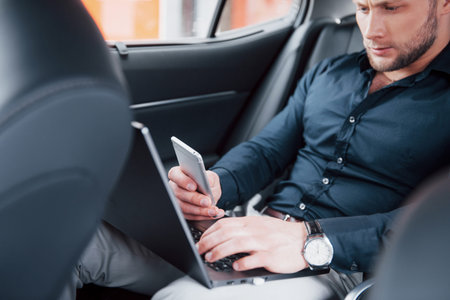 A young businessman working on laptop and talking on the phone while sitting in the cars back. Works...