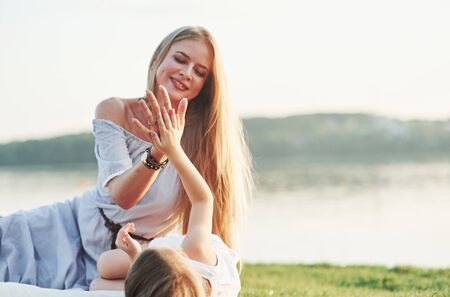 Give me high five. Photo of young mother and her daughter having good time on the green grass with lake at background.の写真素材