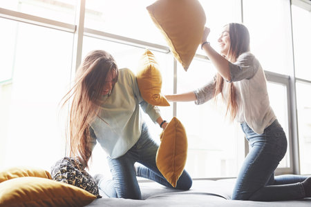 Two beautiful young twins sisters spending time together and are battling with pillows. Siblings...
