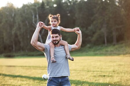 You will grow up like me in the future, but now let me take you on my shoulders. Photo of dad with his daughter at beautiful grass and woods at background.の写真素材