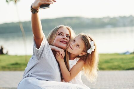 Lets take a selfie. Photo of young mother and her daughter having good time on the green grass with lake at background.の写真素材