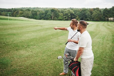 Two stylish men holding bags with clubs and walking on golf course.の写真素材