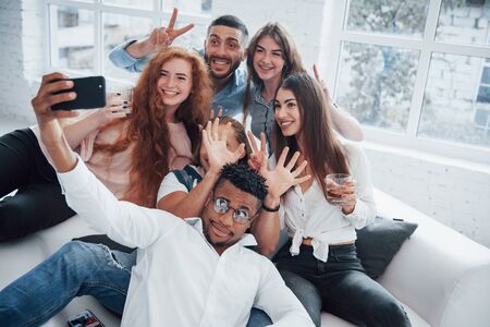 Taking a picture. Cheerful young friends having fun and drinking in the white interior.の写真素材