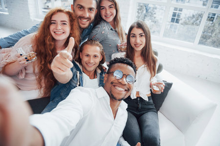 Thumb up. Cheerful young friends having fun and drinking in the white interior.