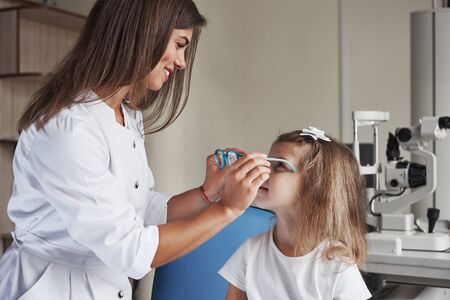 Smiling woman selects glasses. Little girl tries new blue glasses in ophthalmology office with female doctor.の写真素材