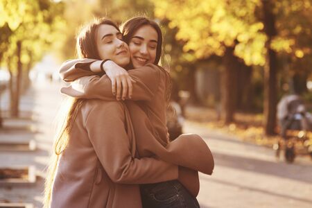 Side profile of young pretty smiling brunette twin girls hugging and having fun in casual coat standing close to each other at autumn sunny park alley on blurry background.の写真素材