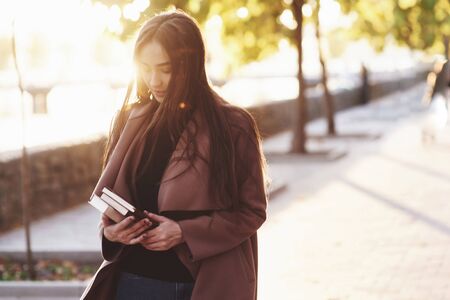 Young pretty brunette girl standing, holding stack of books in hands with her look down, eyes closed, wearing casual coat and walking at autumn sunny park alley on blurry background with a copy space.の写真素材