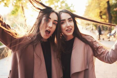 Close portrait of two cheerful and pretty, young brunette twin girls in casual coat having fun and playing with hair outdoor in a sunny autumn park on the blurry background.の写真素材