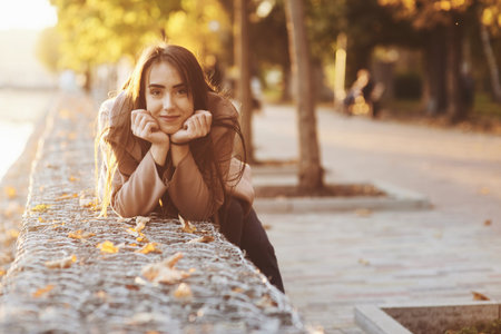 Young, pretty brunette girl leaned on the wall with her two hands under the chin, wearing casual coat with an autumn sunny park on the blurry background with a copy space.の写真素材