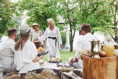 Women walks to the table. Group of adult friends have a rest and conversation in the backyard of...