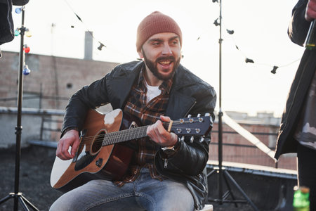 Young bearded man. Guy with acoustic guitar sings. Friends have fun at rooftop party with decorative...