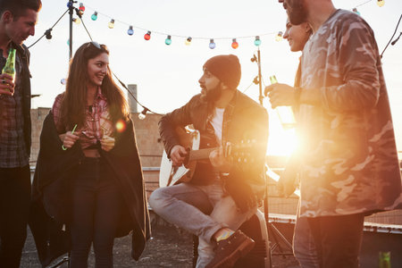Girl is enchanted. Guitarist sits and singing for his friends at the rooftop with decorative colored...