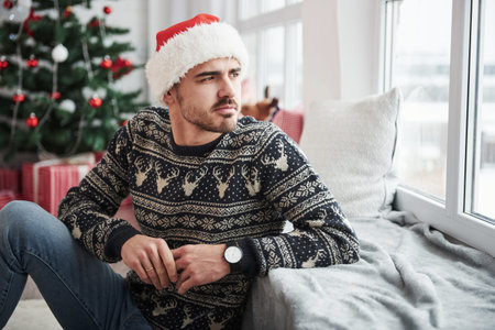 Leaning on the window sill. Photo of man in santa hat and holiday clothes looks through the window....