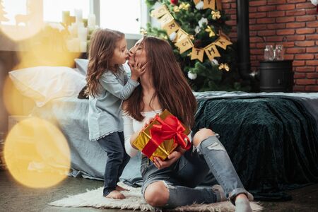 Cute scene of kiss. Mother and daughter sits in holiday decorated room and holds gift box.の写真素材