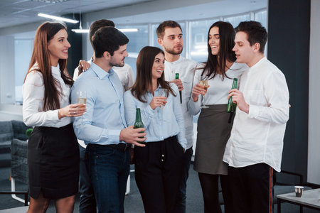 Drunk guy telling something interesting. Everyone is listening. Photo of young team in classical clothes holding drinks in the modern good lighted office.