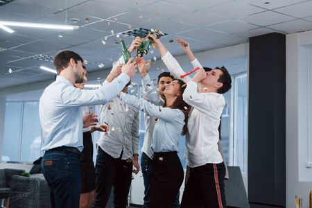 Hands up. Photo of young team in classical clothes celebrating success while holding drinks in the modern good lighted office.の写真素材