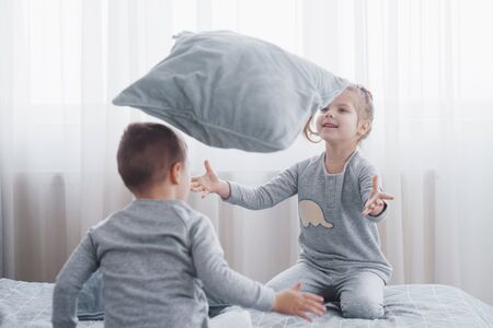 Happy kids playing in white bedroom. Little boy and girl, brother and sister play on the bed wearing pajamas. Nursery interior for children. Nightwear and bedding for baby and toddler. Family at homeの写真素材