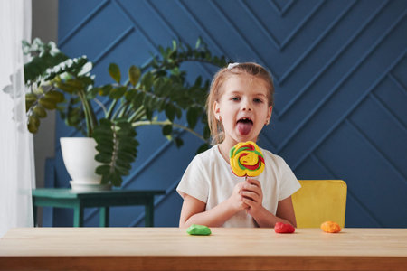 Shows her crafted candy. Children playing with colored plasticine on the wooden table at home.