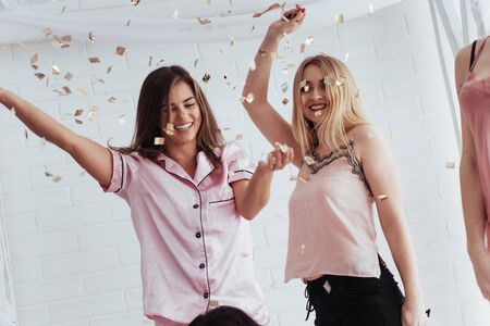 Emotional faces. Confetti in the air. Young girls have fun on the white bed in nice room.の写真素材