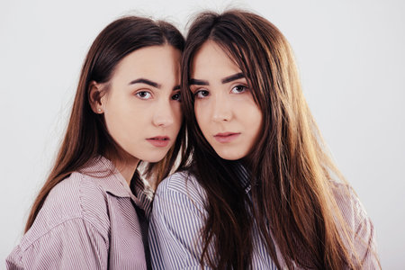 Cute girls looking into the camera. Two sisters twins standing and posing in the studio with white...